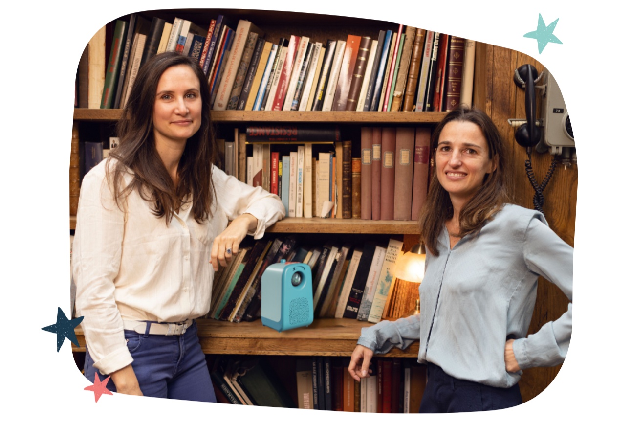 Laurène Altmayer and Aliénor Bouvier-Lewi standing beside a turquoise Tikino projector in front of bookshelves.