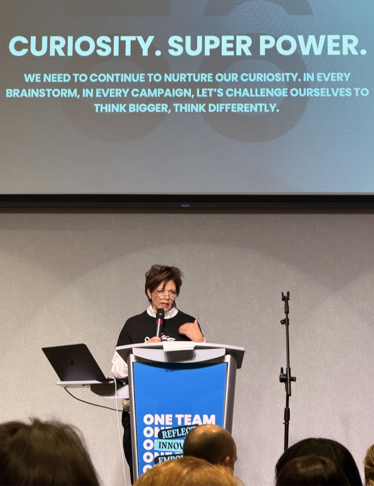 Laurie Fleck speaking at a podium beneath a screen reading Curiosity. Superpower.