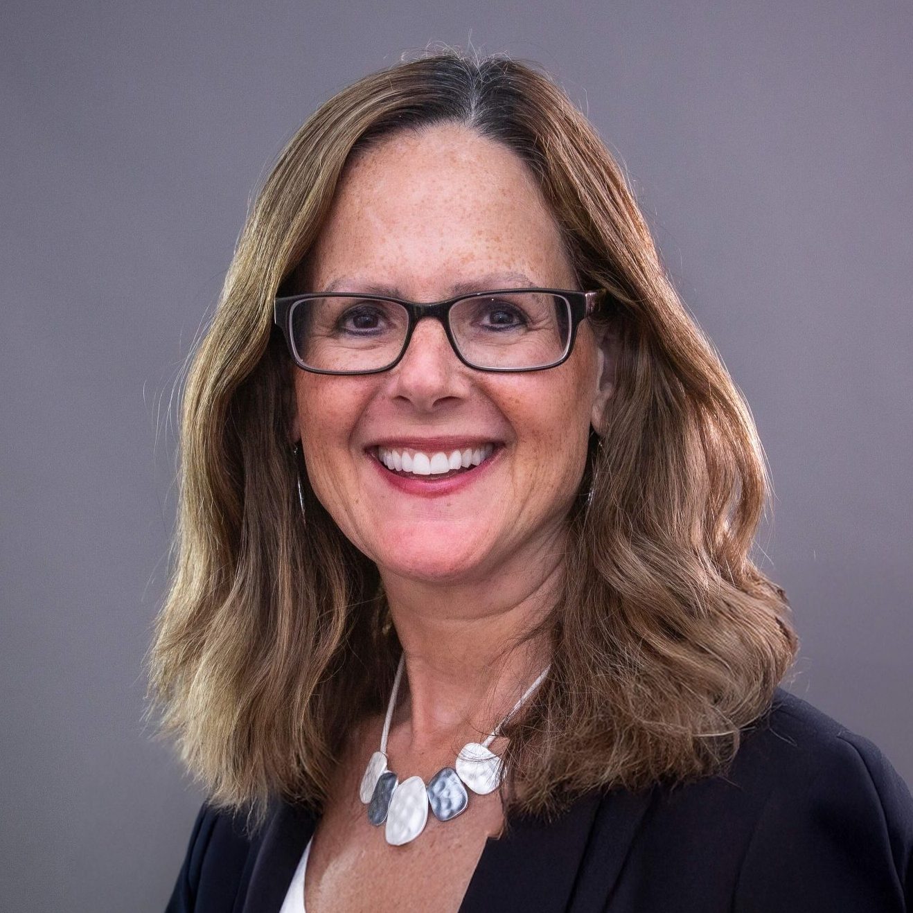 Laurie Klausner smiling in a dark blazer and glasses against a gray studio background