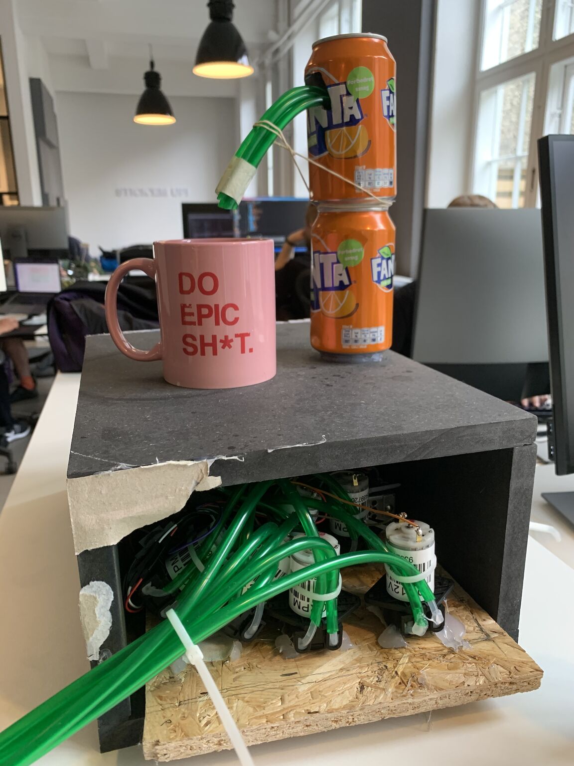 An improvised cocktail machine with green tubing, pumps, two orange cans and a pink mug