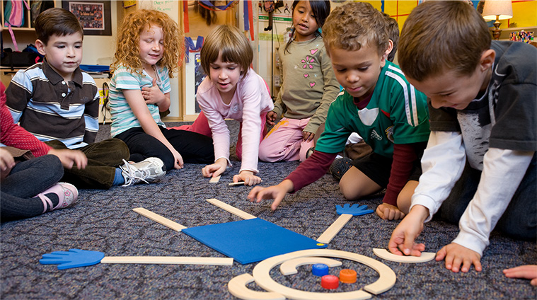Children arrange wooden lines, curves and a blue mat to build Mat Man on a classroom carpet.