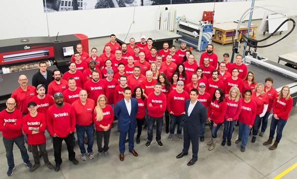 Tectonics staff gathered on the factory floor; Lee Skandalaris stands at the front right in a navy suit.