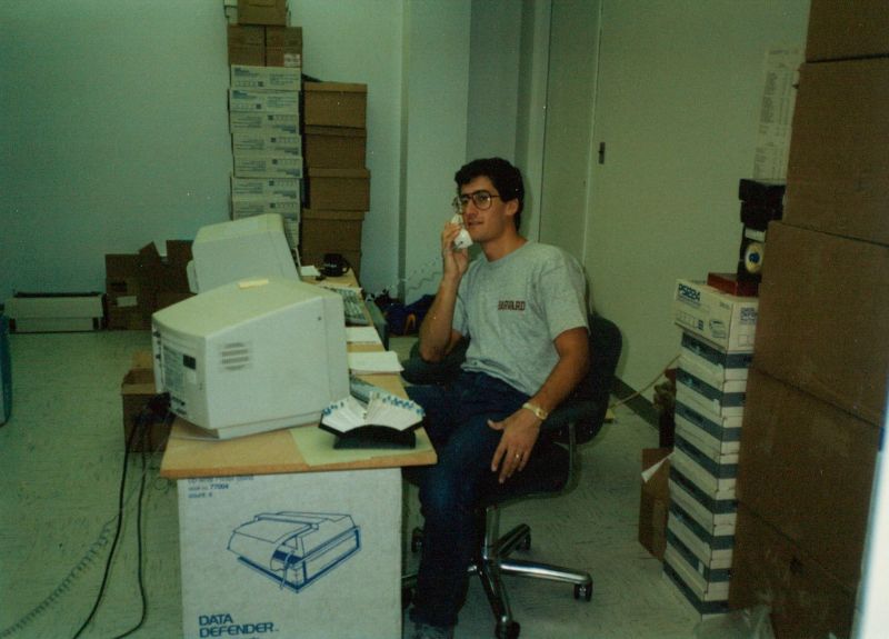 A young Leo Russell wearing glasses and a Harvard T-shirt, talking on a telephone at a desk surrounded by computer boxes.