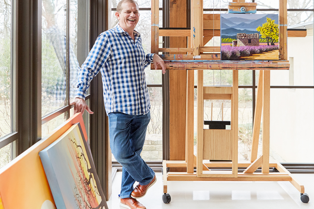 Leon Backes standing in his bright art studio beside a tall easel and colorful oil paintings