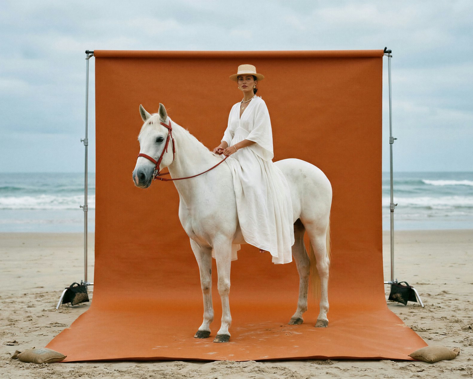 A woman in white riding a white horse in front of an orange studio backdrop on a beach