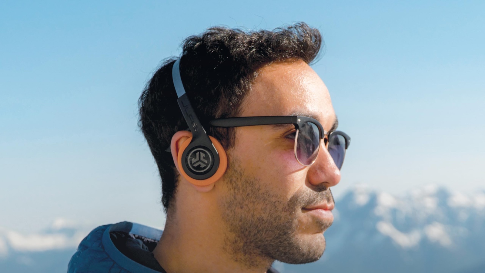 A person wearing JLab open-ear headphones outdoors against a mountain sky