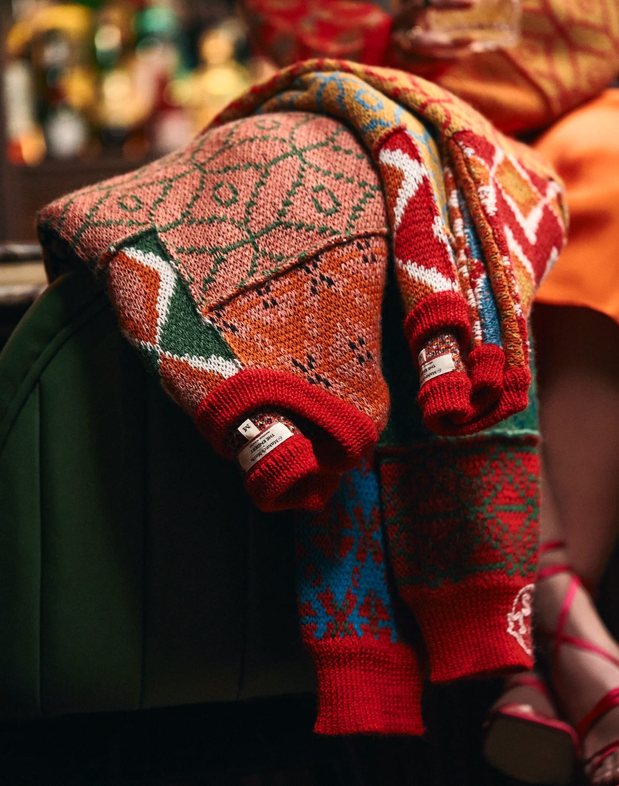 Colorful Maker’s Mark and The Endery knitted holiday sweaters draped over a wooden chair