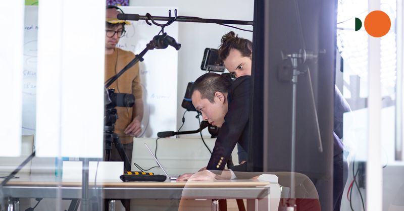Liang Lin leans over a laptop while a camera crew films inside an office.