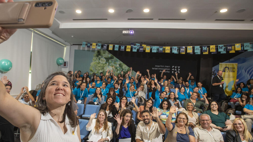 Lili Ben Ami holds up her phone for a selfie as a room of Michal Sela hackathon participants waves behind her.