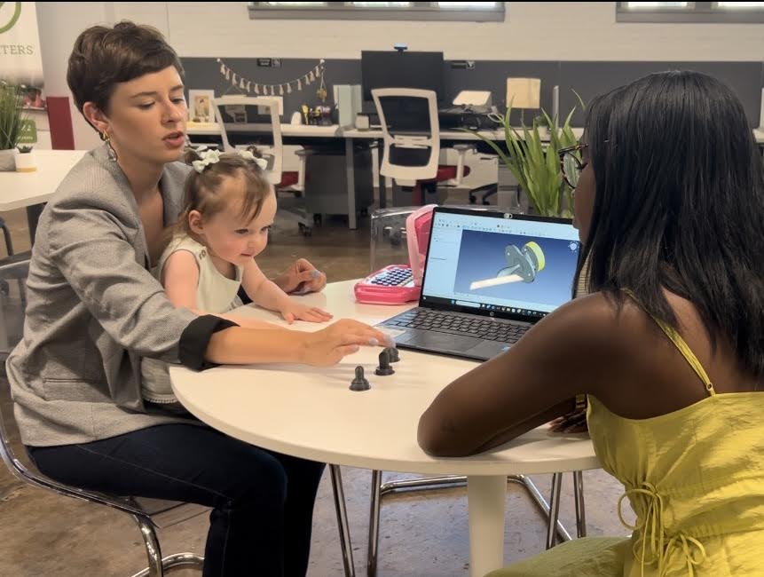 Lindsay deOliveira at a worktable with her daughter, product components and a pacifier model on a laptop