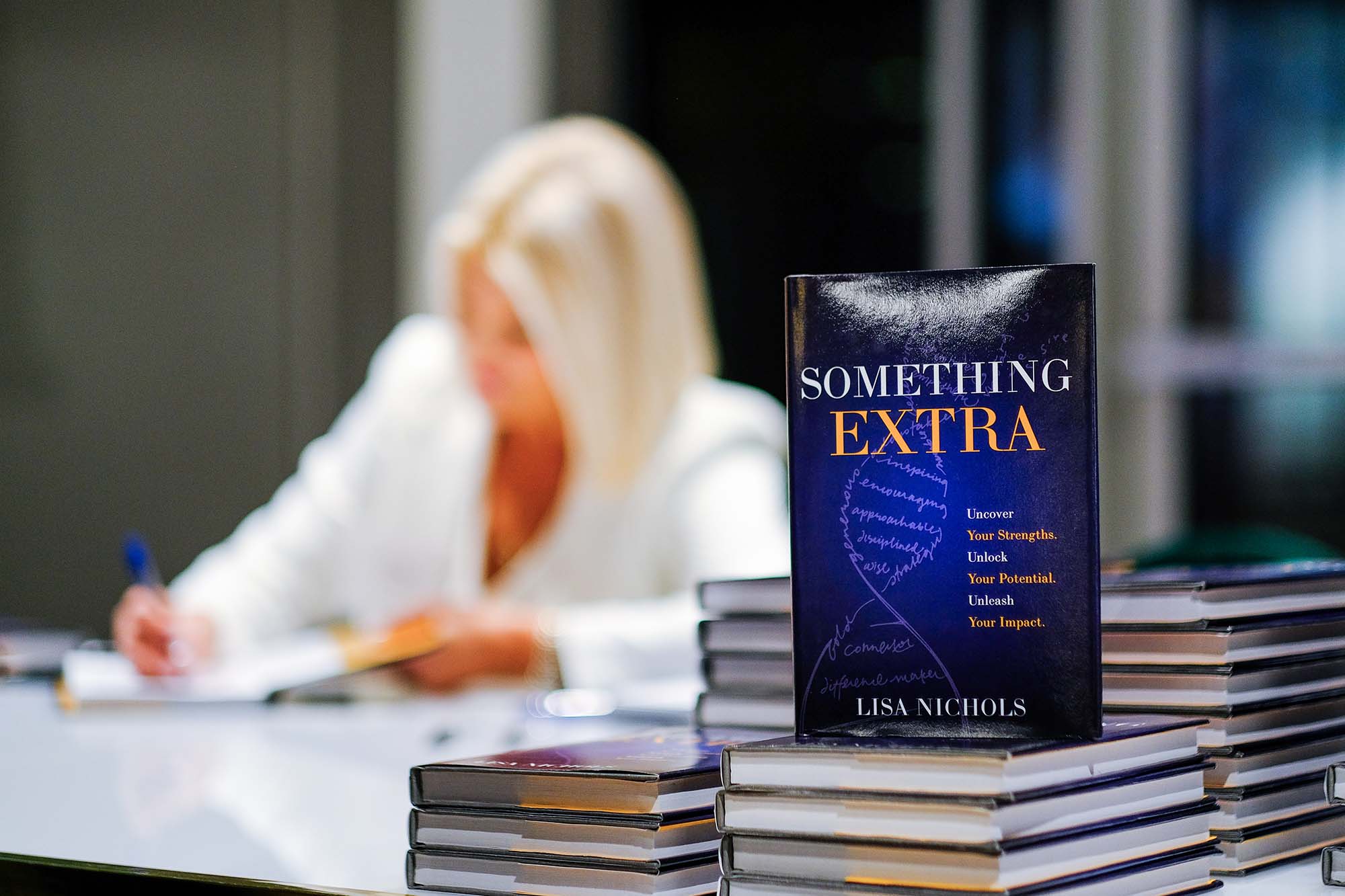 Copies of Something Extra in the foreground while Lisa Nichols signs books behind them