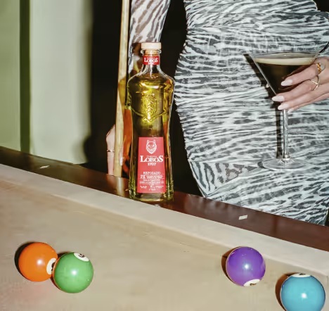 A Lobos 1707 Reposado bottle beside a cocktail and colorful pool balls