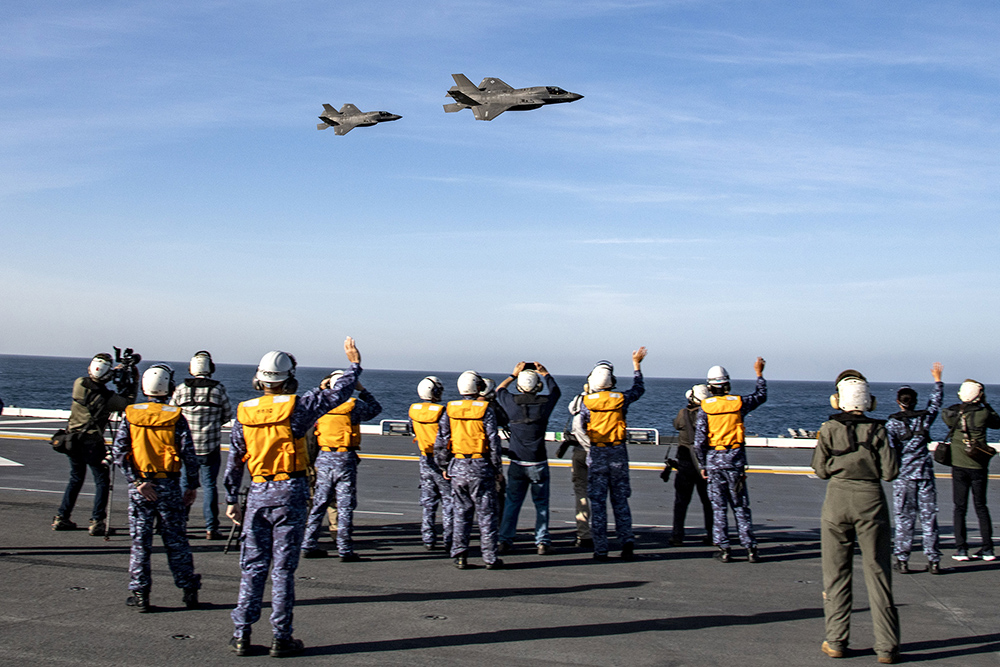 Flight-deck personnel watch two F-35 aircraft flying over a ship