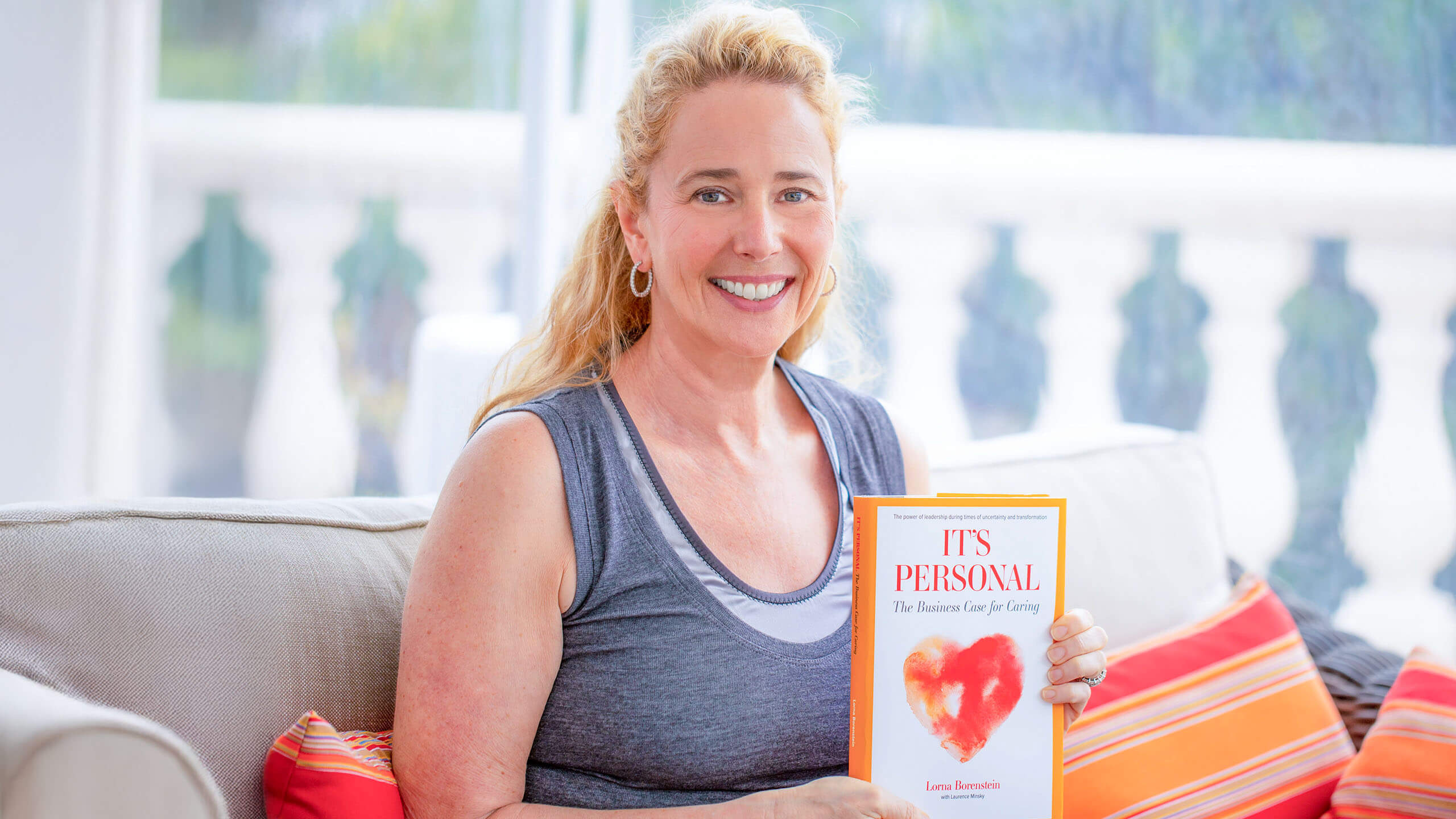 Lorna Borenstein seated on a sofa, holding It’s Personal: The Business Case for Caring