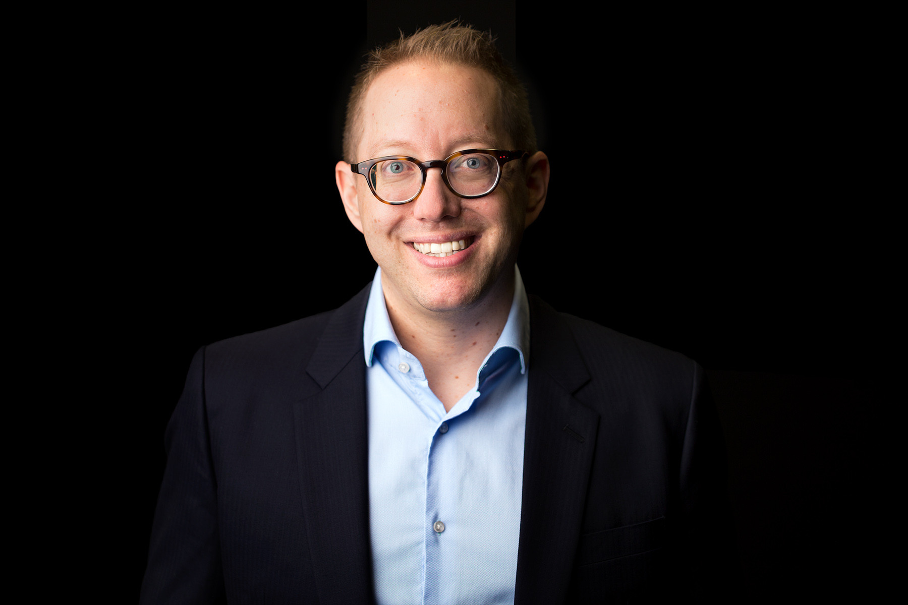 Louis Adam in a dark jacket and light blue shirt against a black background