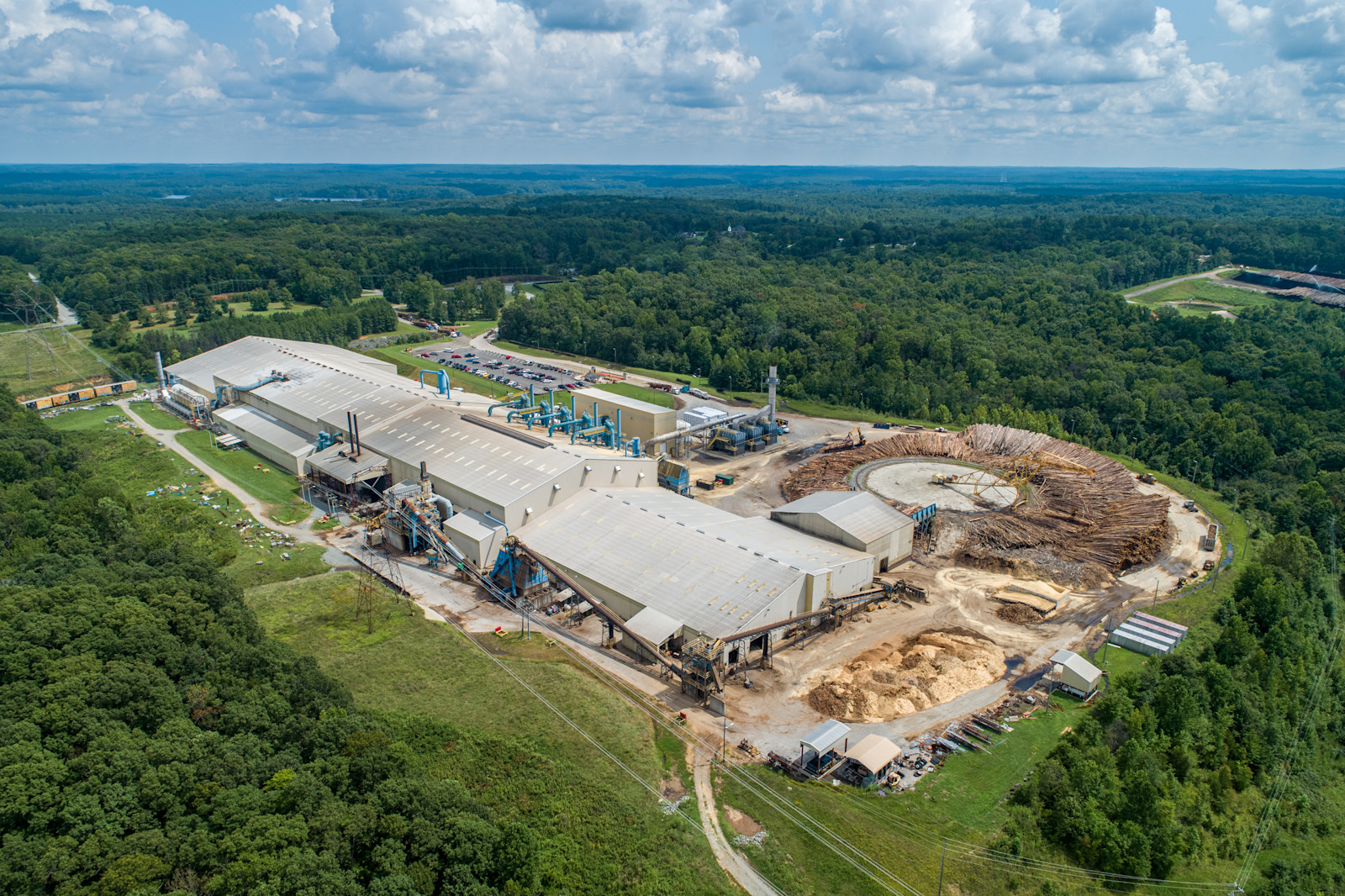 Aerial view of an LP manufacturing facility surrounded by forest, with logs stacked beside the plant