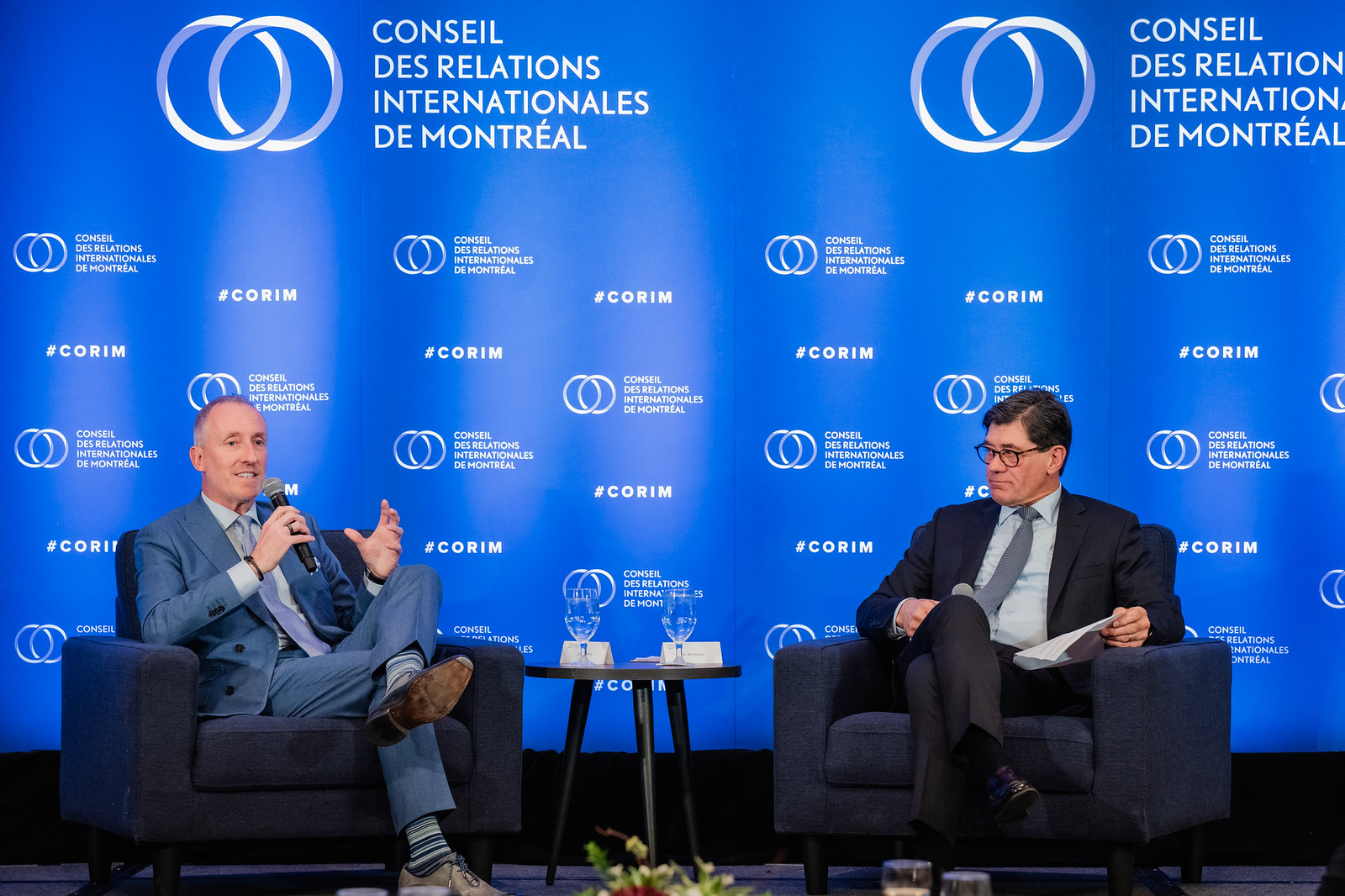 Luc Fortin speaks into a microphone while Luc Bertrand listens on stage at CORIM.