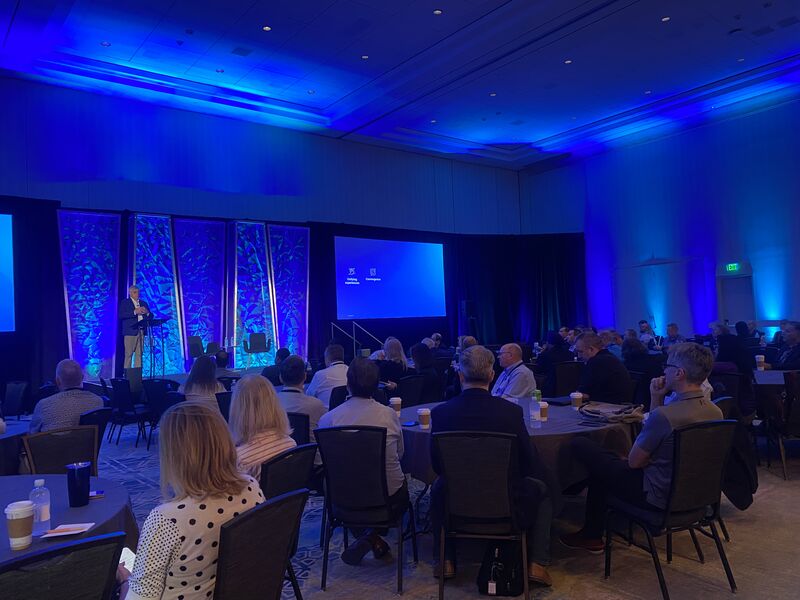 Audience seated in a blue-lit conference room as a speaker presents on stage