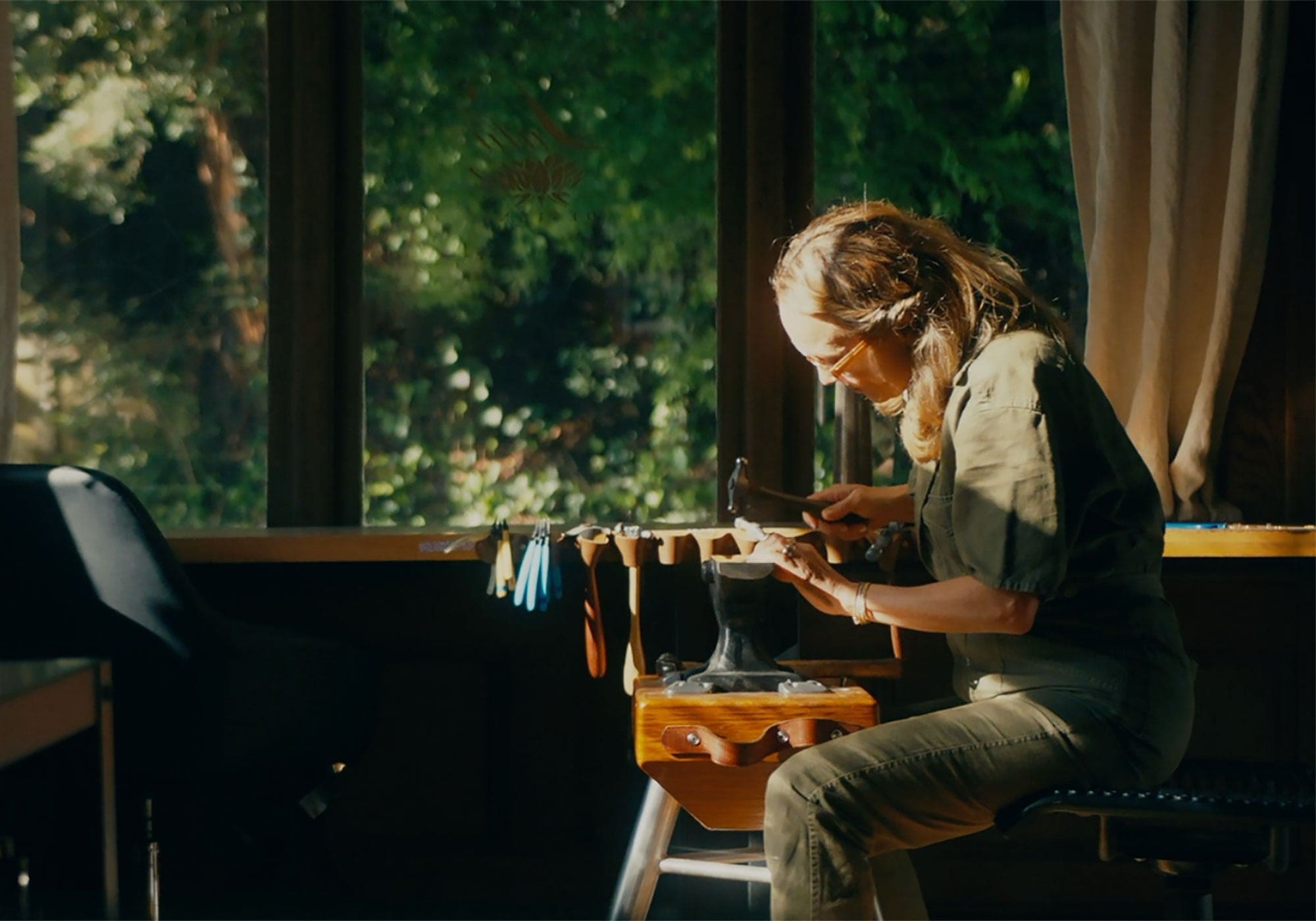A Lulu Designs jeweler working with a hammer beside a sunlit studio window