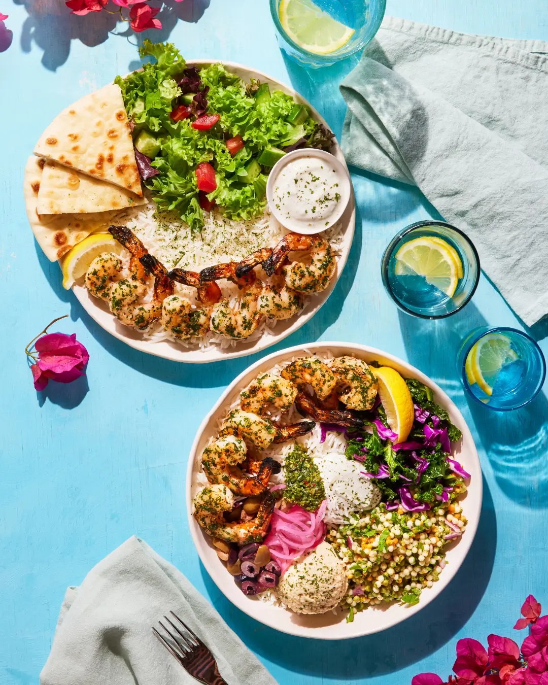 Two Luna Grill shrimp meals with rice, pita, greens, hummus and colorful vegetables on a blue table