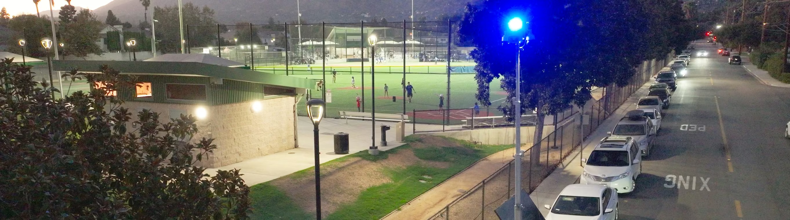 An LVT surveillance mast with a blue light beside a busy sports park at dusk