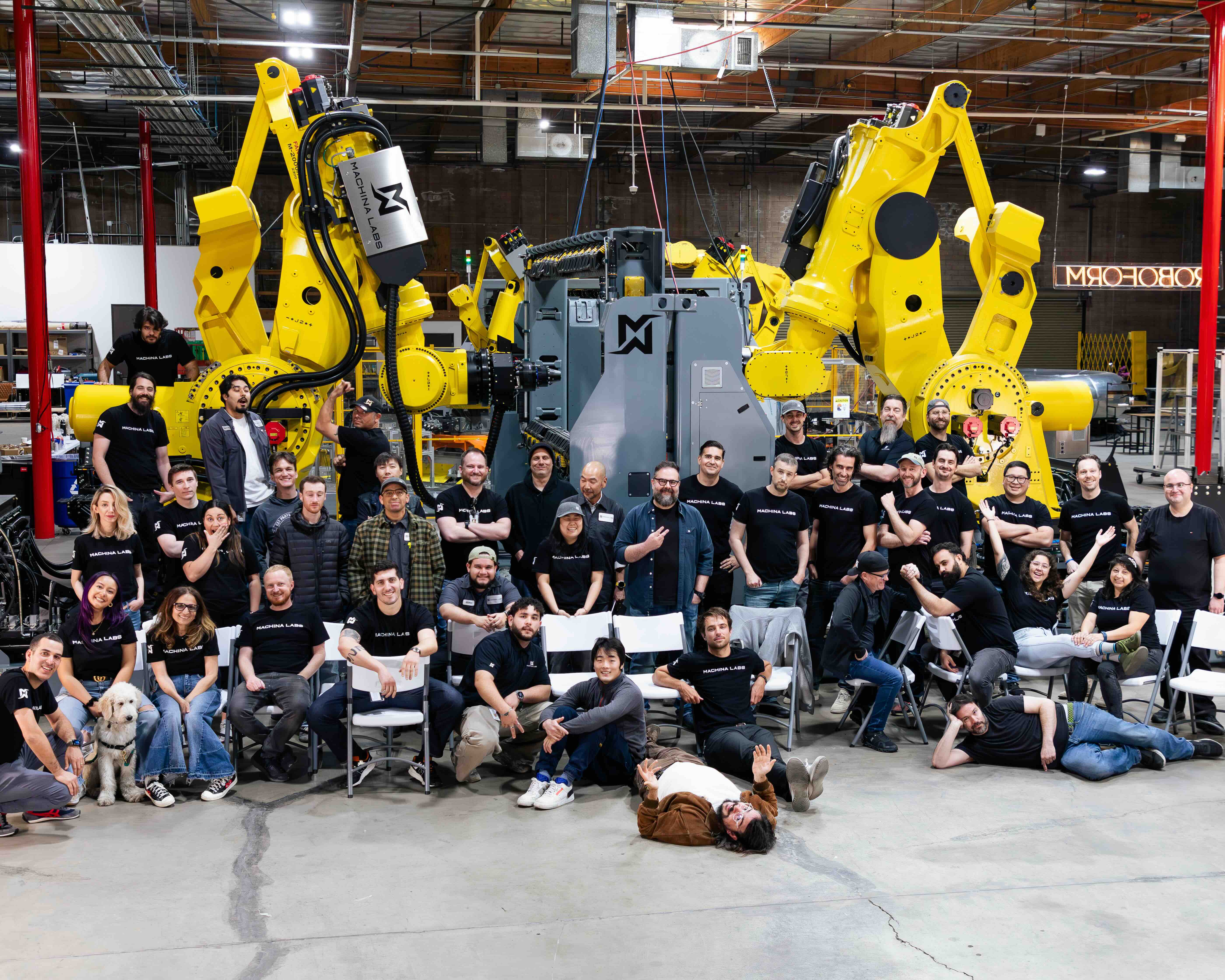 Machina Labs team gathered in the workshop beside yellow industrial robots