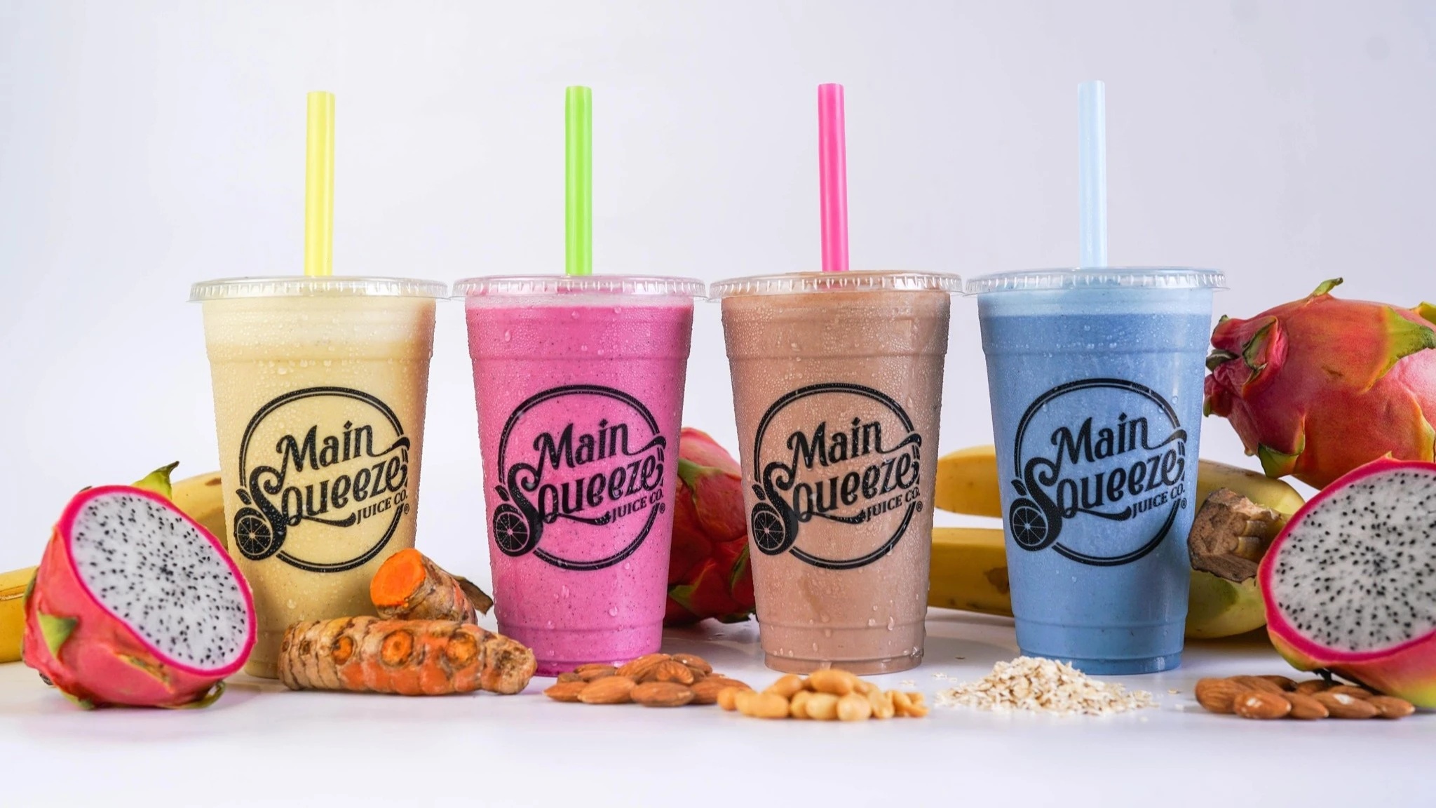 Four Main Squeeze smoothies in branded cups surrounded by fruit, nuts, and other ingredients