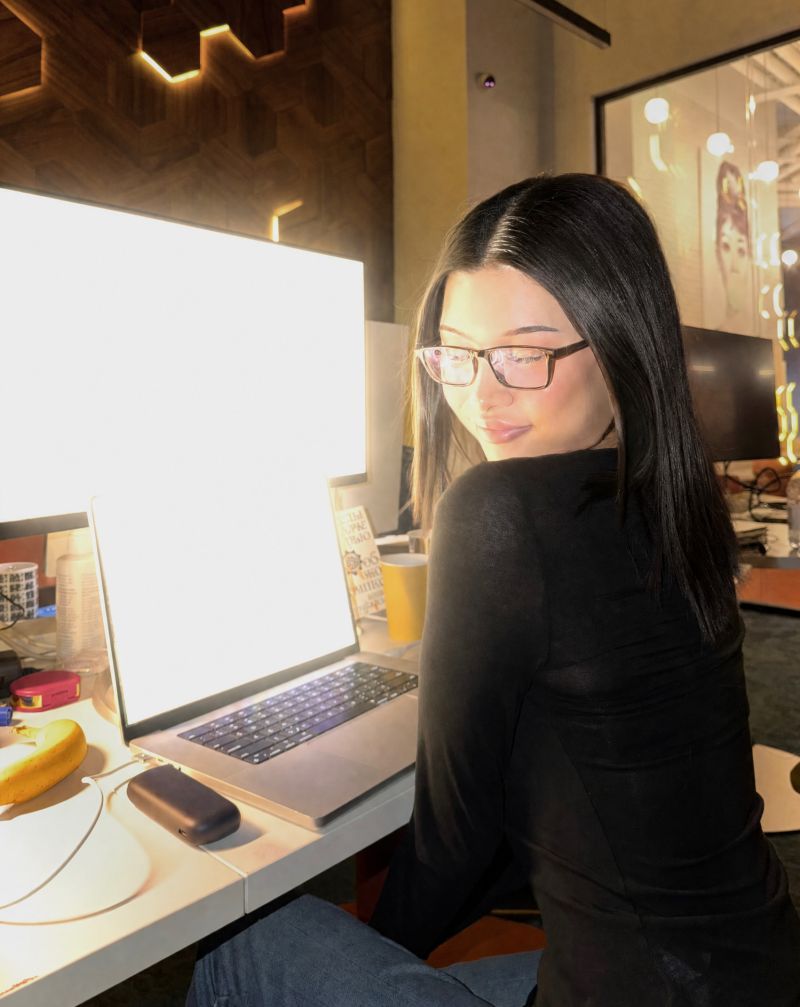 Maira Alyakbar seated beside a brightly lit laptop in a workspace