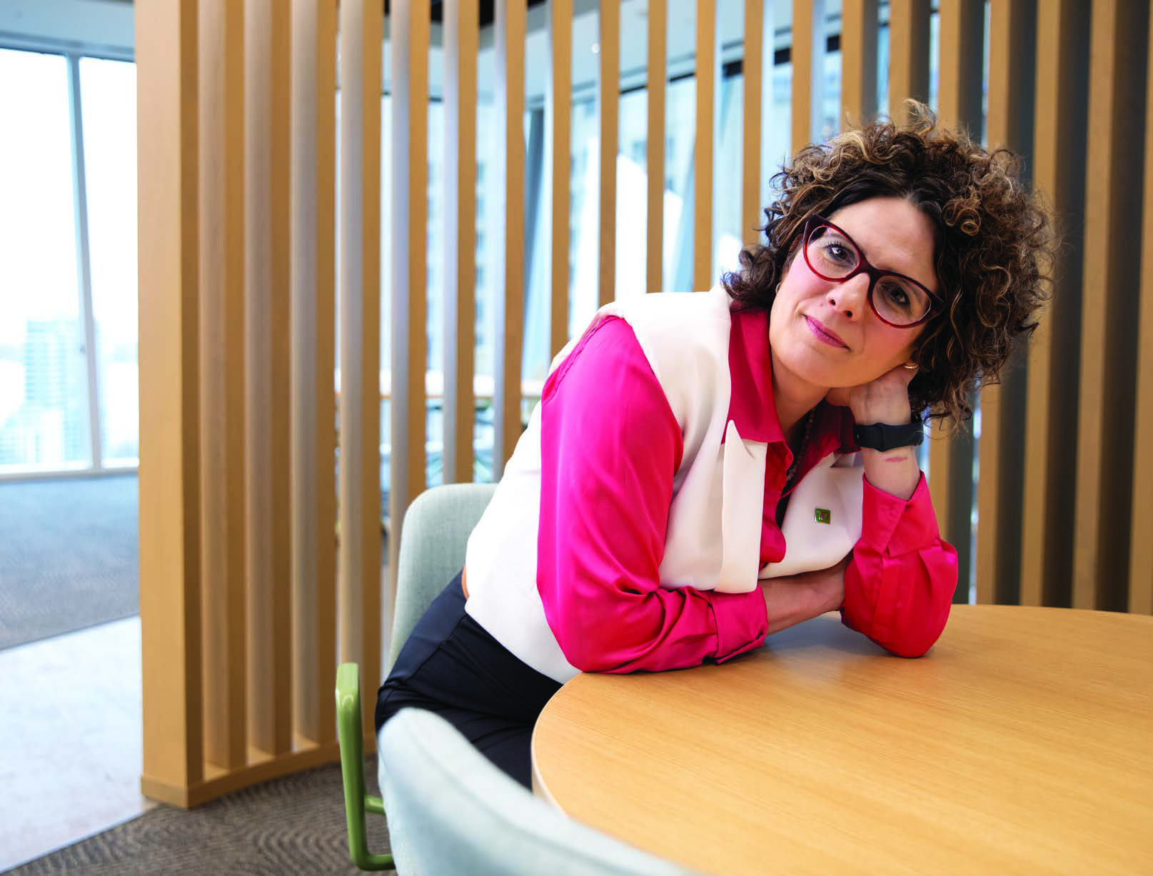 Maja Neable seated at a round table in a bright office, wearing red glasses and a pink shirt