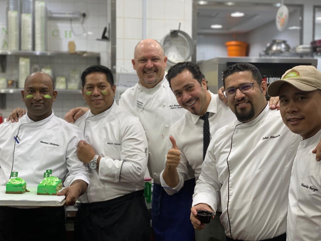 Majed Al Mehdi, in a white shirt and dark tie, smiles with a group of chefs in a kitchen