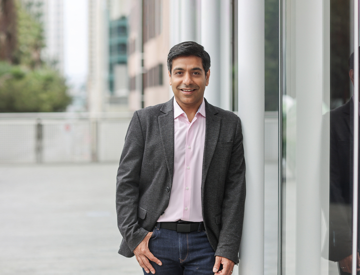 Manav Khurana standing beside a glass wall in a gray blazer and pink shirt