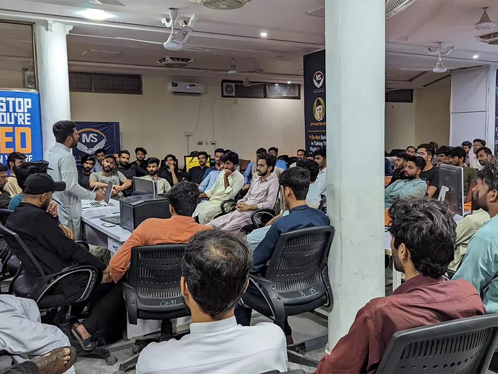 A Manisofts office gathering, with staff seated around computer desks listening to a speaker.