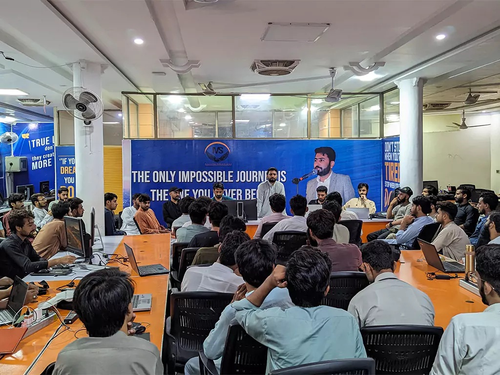 Manisofts staff gathered among desks and laptops in an office, facing a speaker.