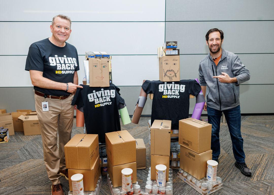 Marc Brown, at left, smiling beside two cardboard figures in HD Supply shirts and a display of collected food.