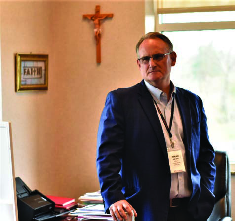Marc Hawk standing in his Granville office, with a crucifix and framed Faith sign on the wall