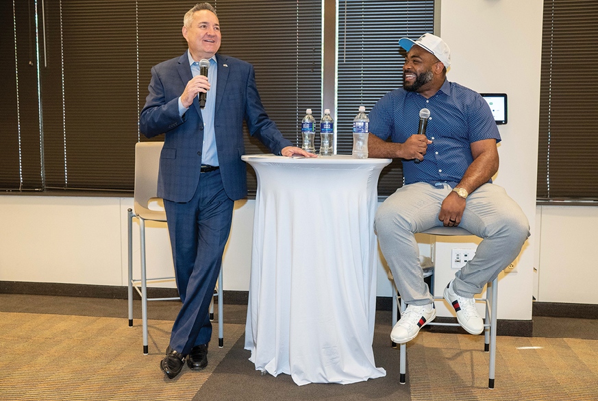 Marc Miller, standing with a microphone, speaks alongside Brandon Graham at an employee town hall.