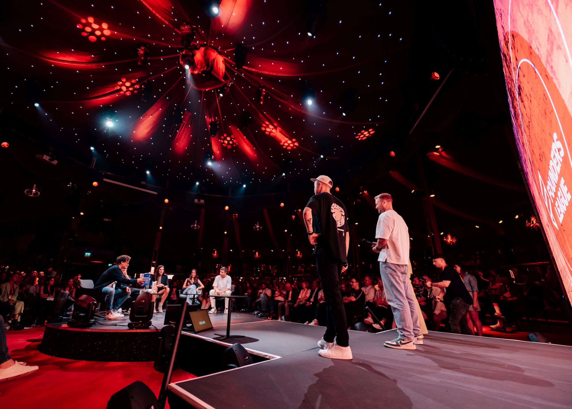 A FOUNDERS LEAGUE pitch show beneath red lighting, with presenters facing a seated jury and audience