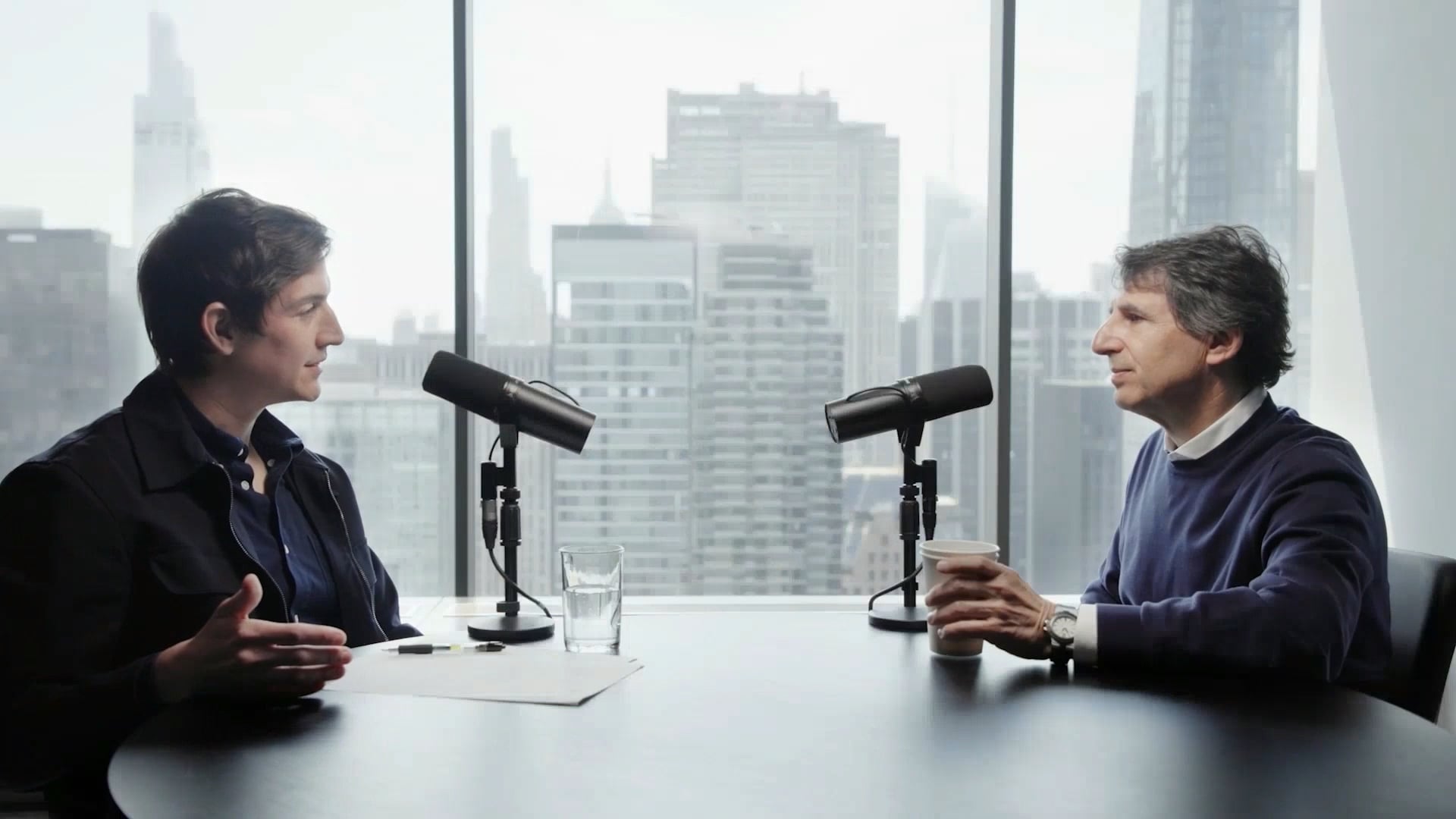 David Haber, left, and Marc Rowan, right, talking across a table with microphones in front of a city skyline