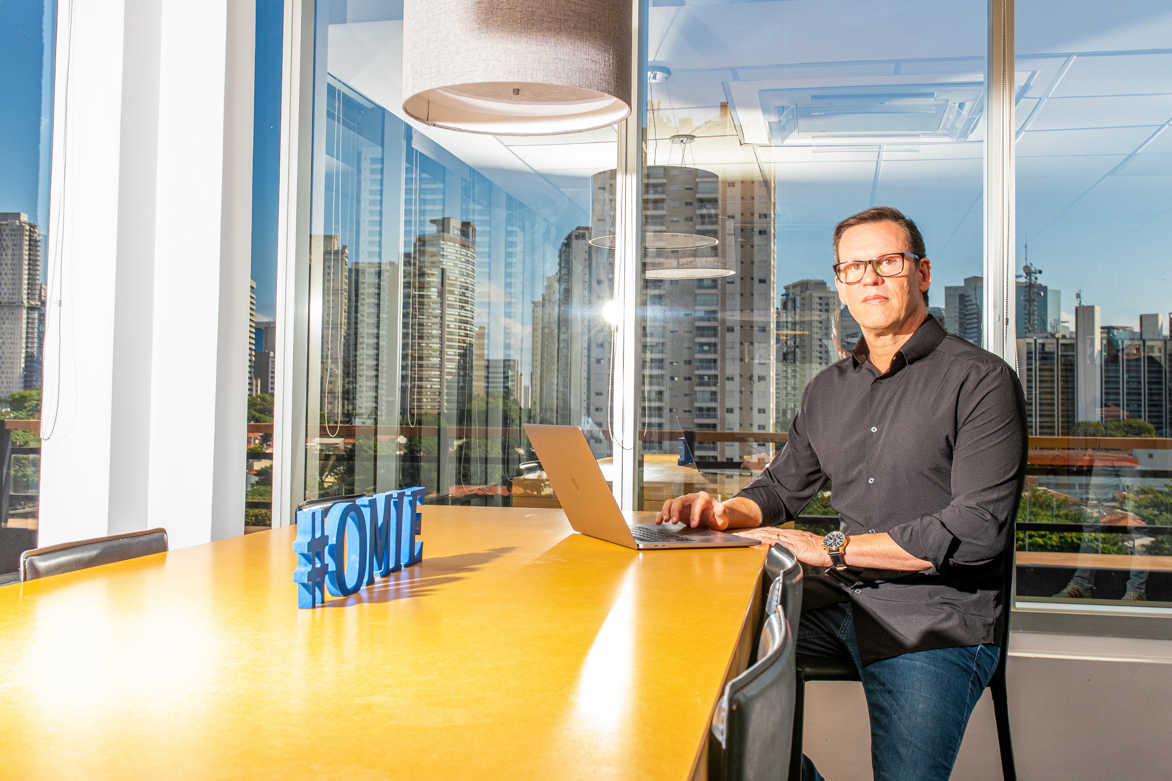 Marcelo Lombardo seated beside a laptop in an Omie office, with São Paulo buildings visible through the windows