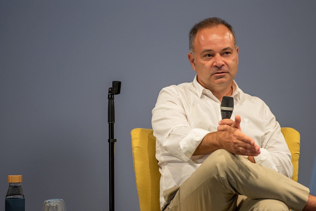 Marco Veremis holding a microphone while seated in a yellow chair at a Fortune Greece Network discussion