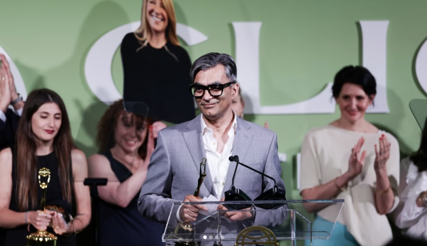Mario Muredda at an awards lectern holding a trophy, surrounded by applauding colleagues