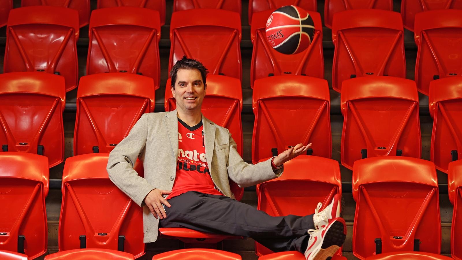 Mark Arena in a Wildcats jersey and grey blazer, tossing a basketball while seated among red stadium seats