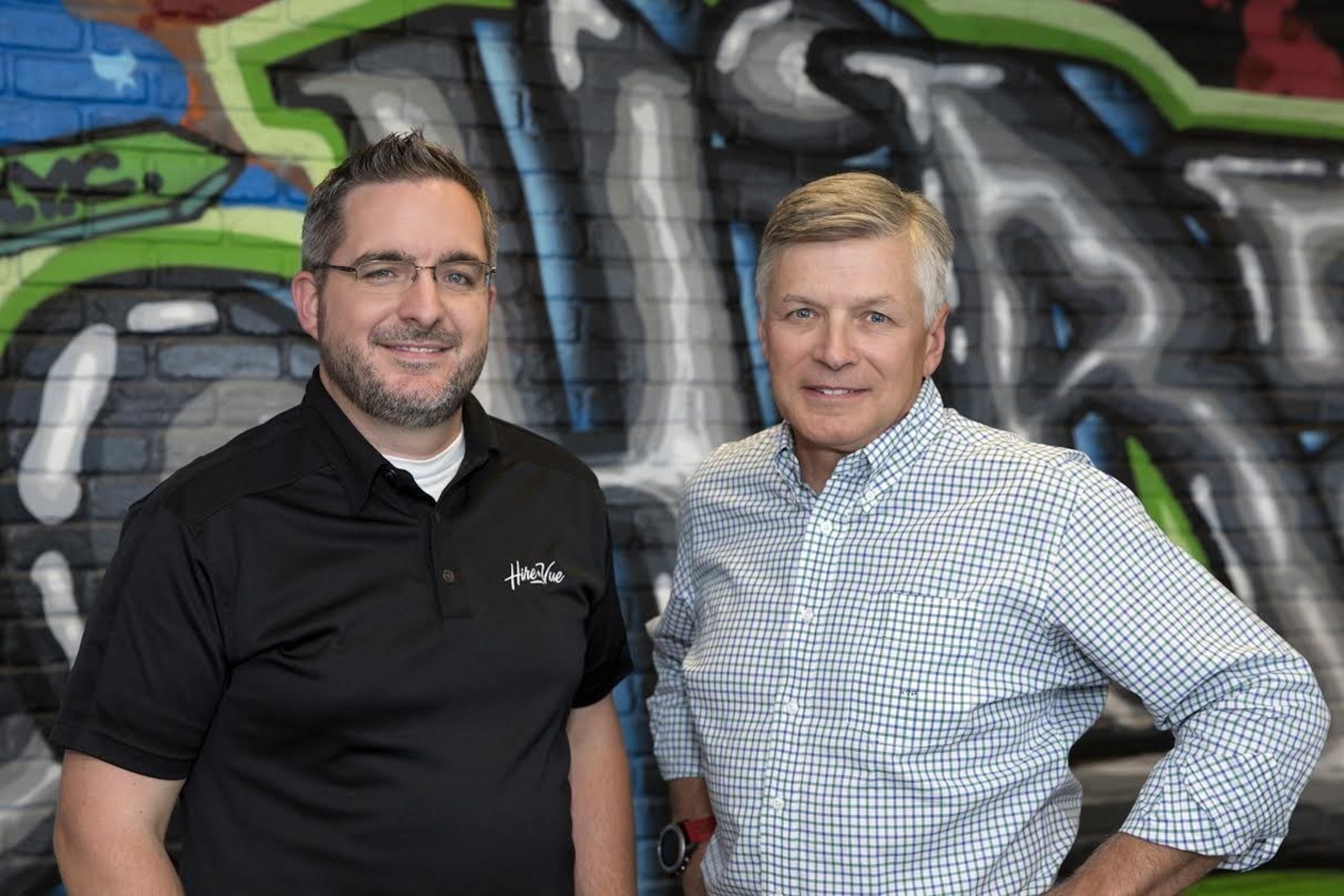 Mark Newman, left, wearing a black HireVue polo beside Kevin Parker in front of a painted wall