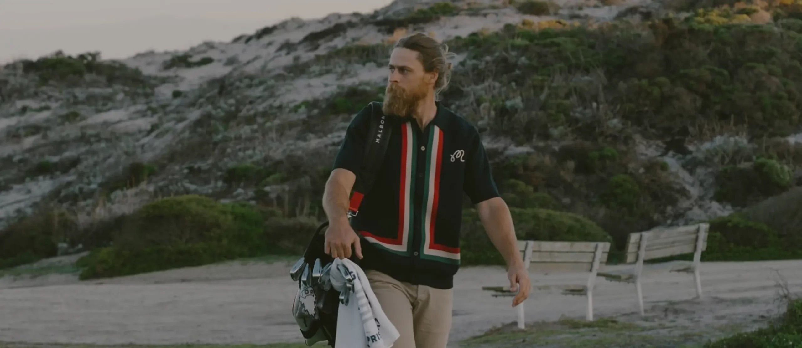 A bearded Malbon golfer in a striped shirt carrying golf clubs beside coastal dunes