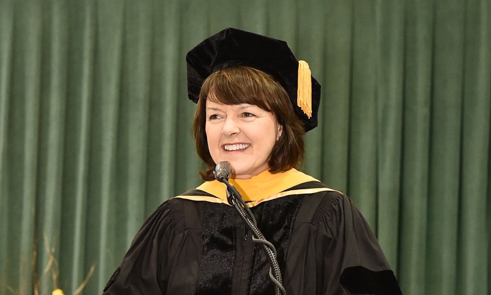 Marnie LaVigne speaking at a microphone in academic robes at SUNY Oswego's December commencement