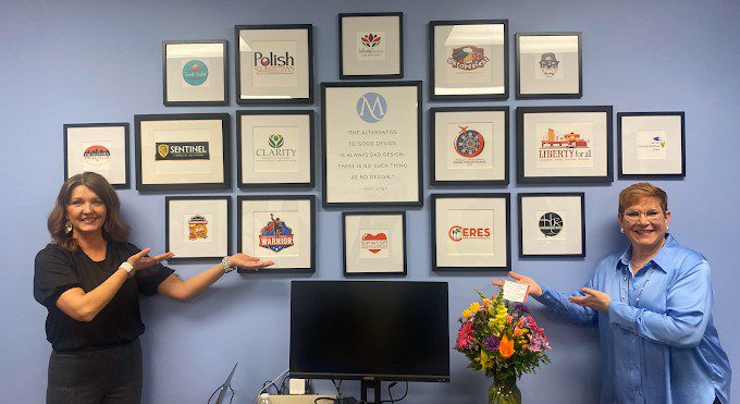 Two MARQUEE team members in their Youngstown office in front of a wall of framed client logos