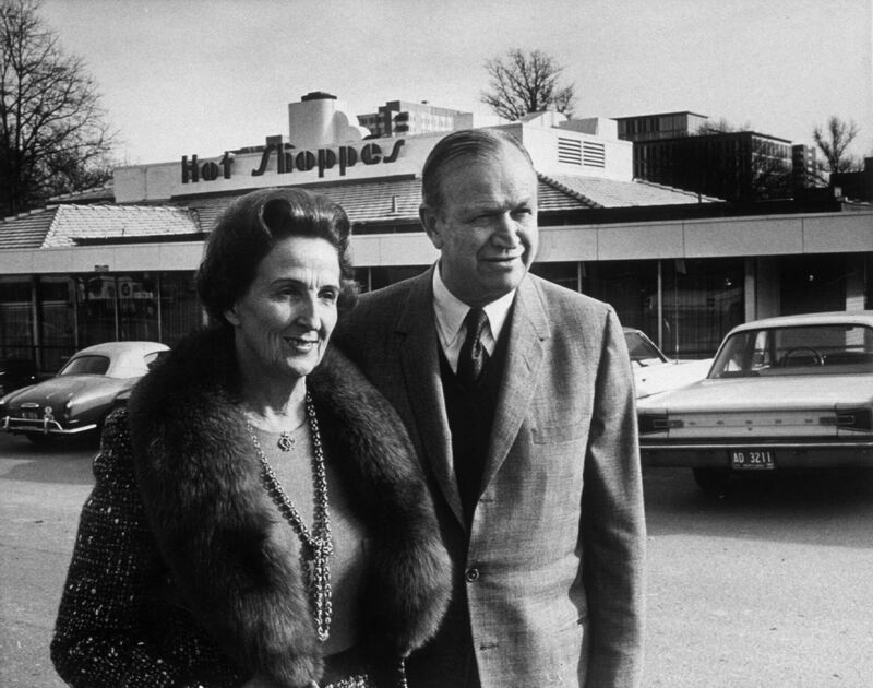 J. Willard and Alice Marriott outside a Hot Shoppes restaurant, with parked cars behind them