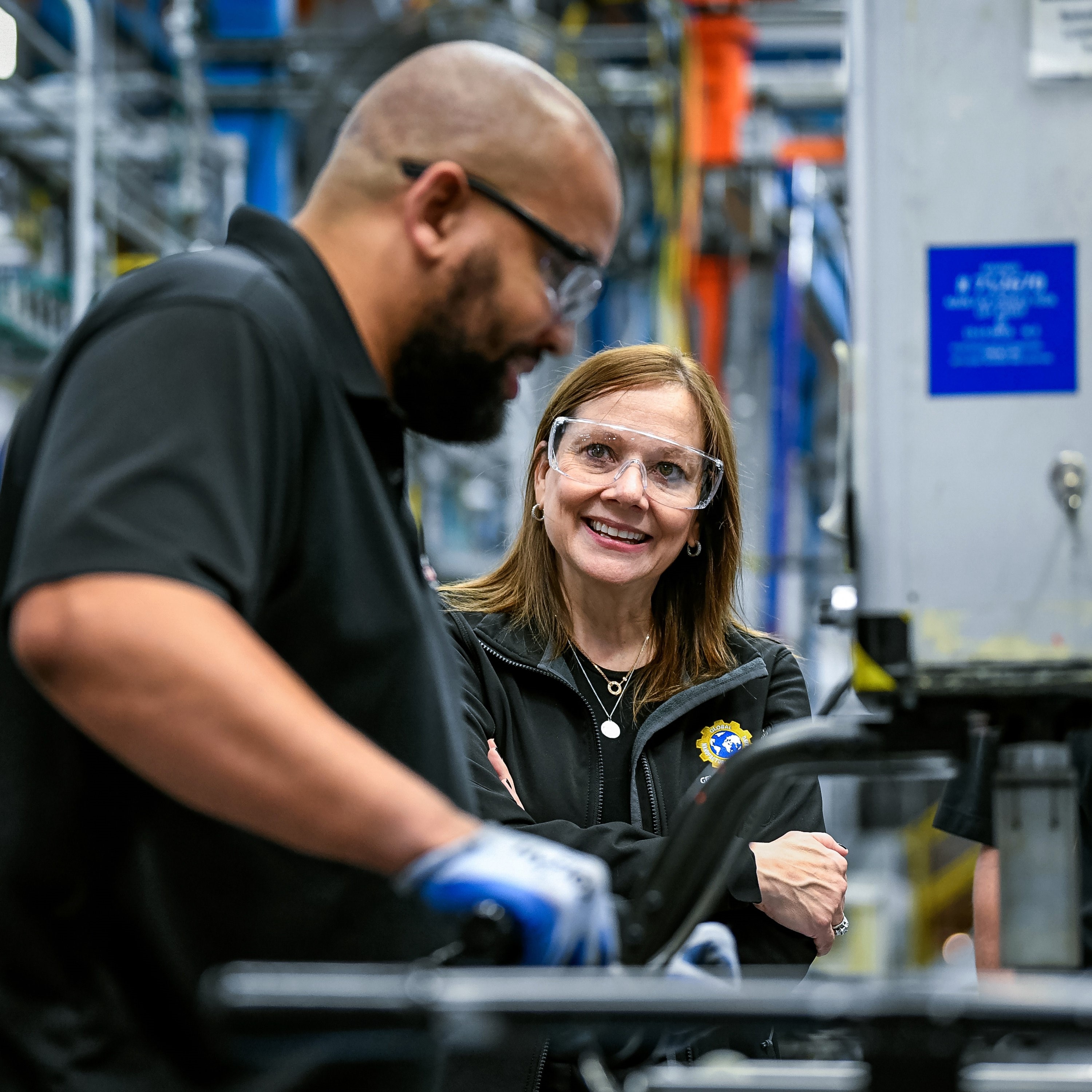 Mary Barra wearing protective glasses, listening to a colleague beside factory equipment.