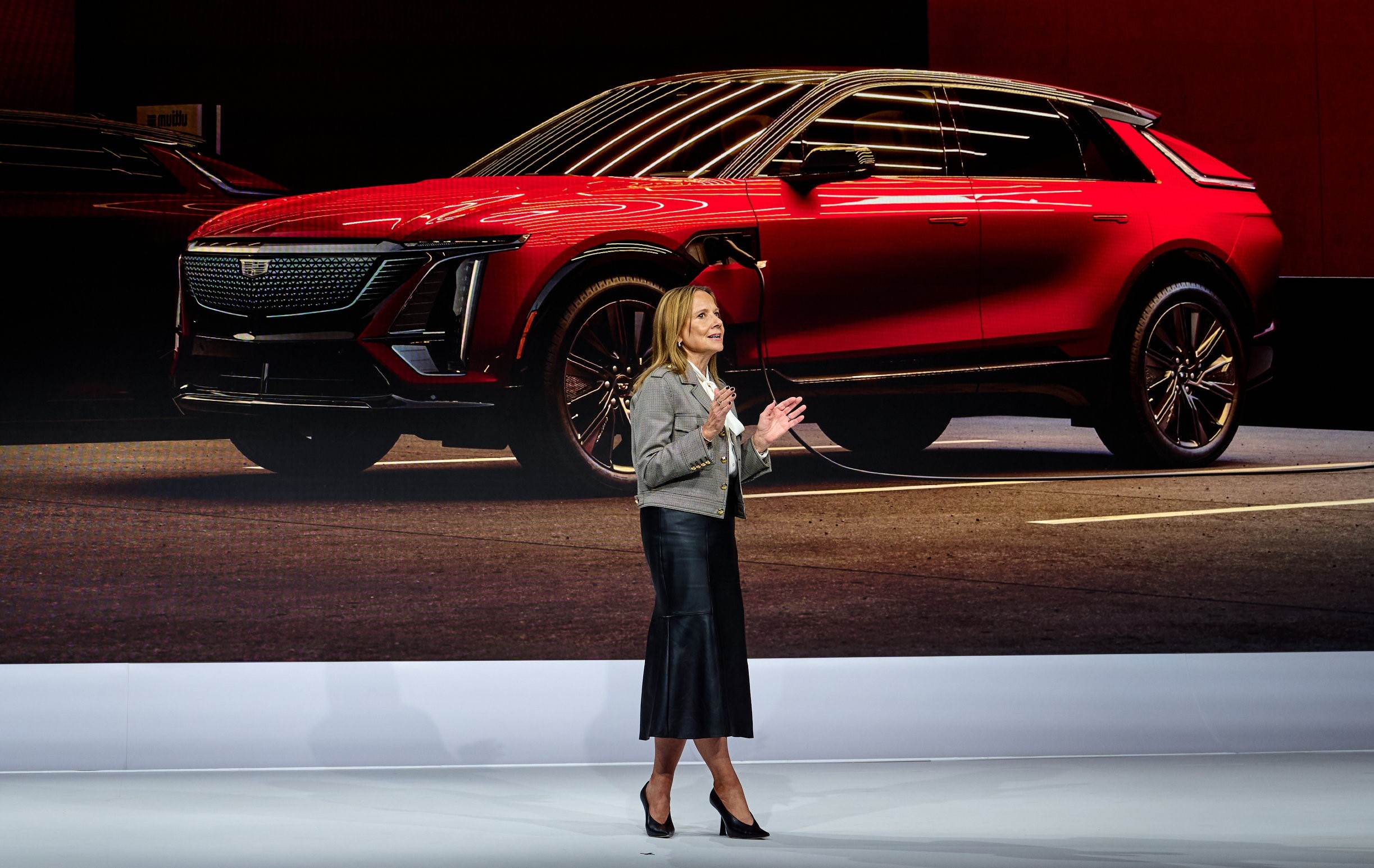 Mary Barra speaks on a stage in front of a large image of a red Cadillac SUV.