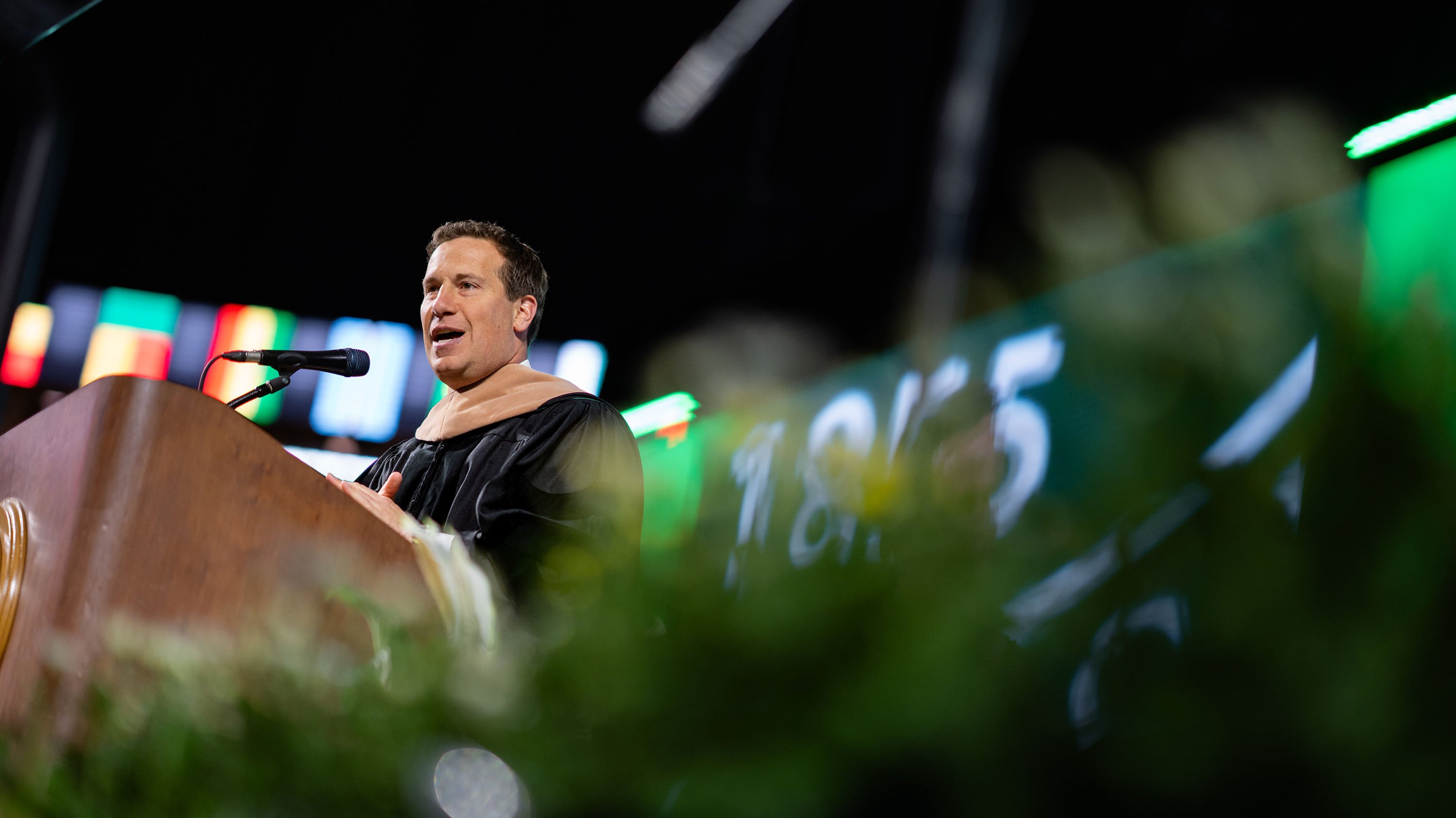 Mat Ishbia in academic robes speaking at a lectern during Michigan State’s 2025 spring convocation